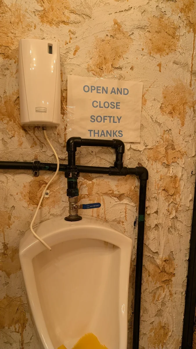 Pisoár s nástenkou s pokynom na jemné zatváranie v rustikálnom prostredí.