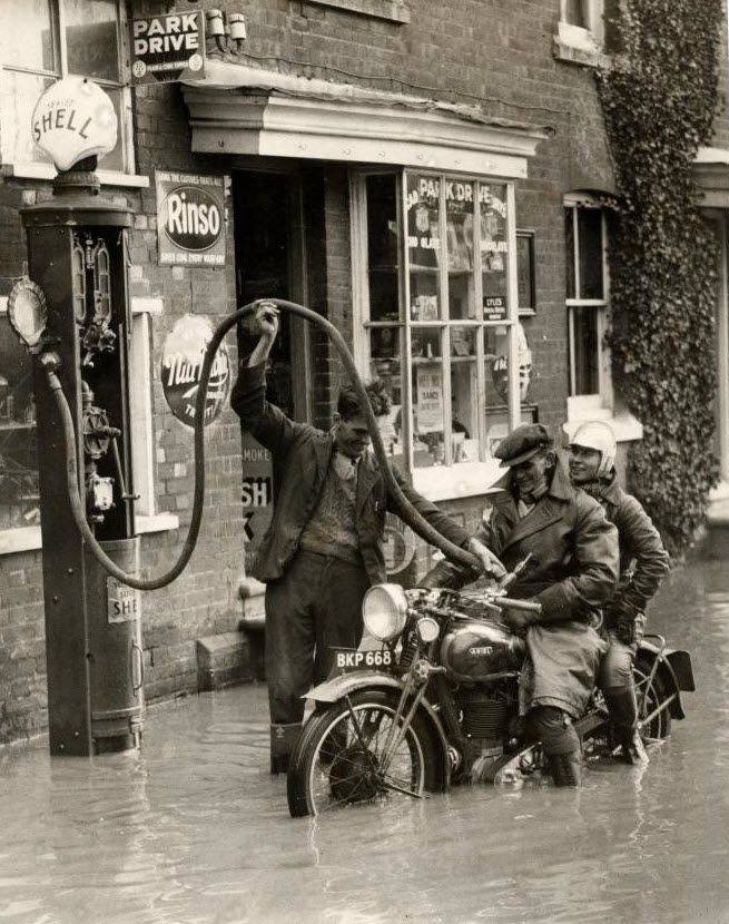 Motocyklisti a obsluha čerpadla sú zaplavení vodou pri historickej benzínovej stanici.
