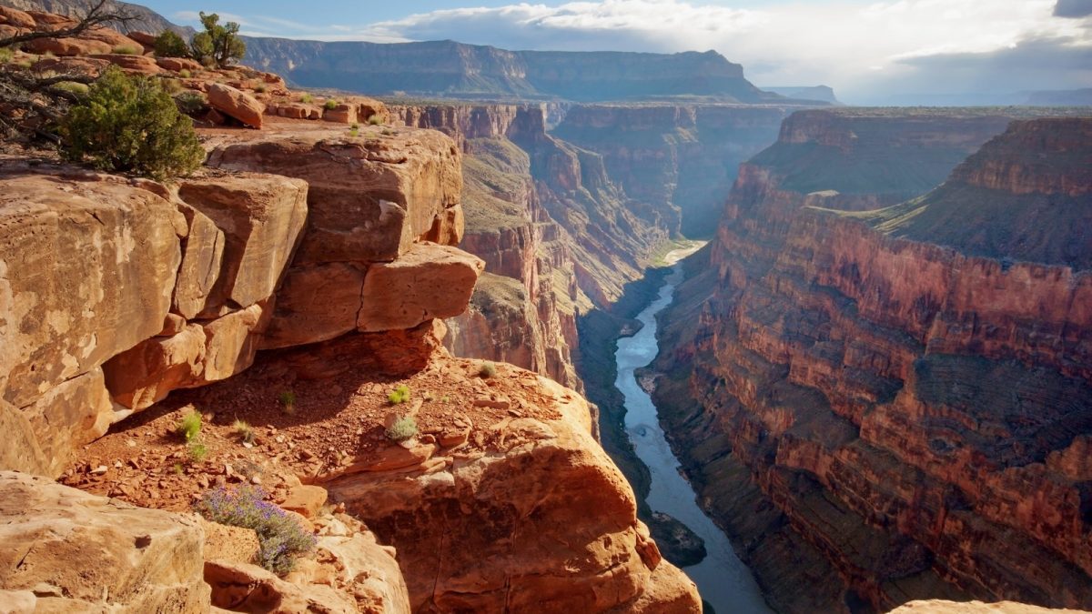 Grand Canyon na slnečnom svetle, s riekou tečúcou v údolí, obklopený strmými červenými skalami a vegetáciou.