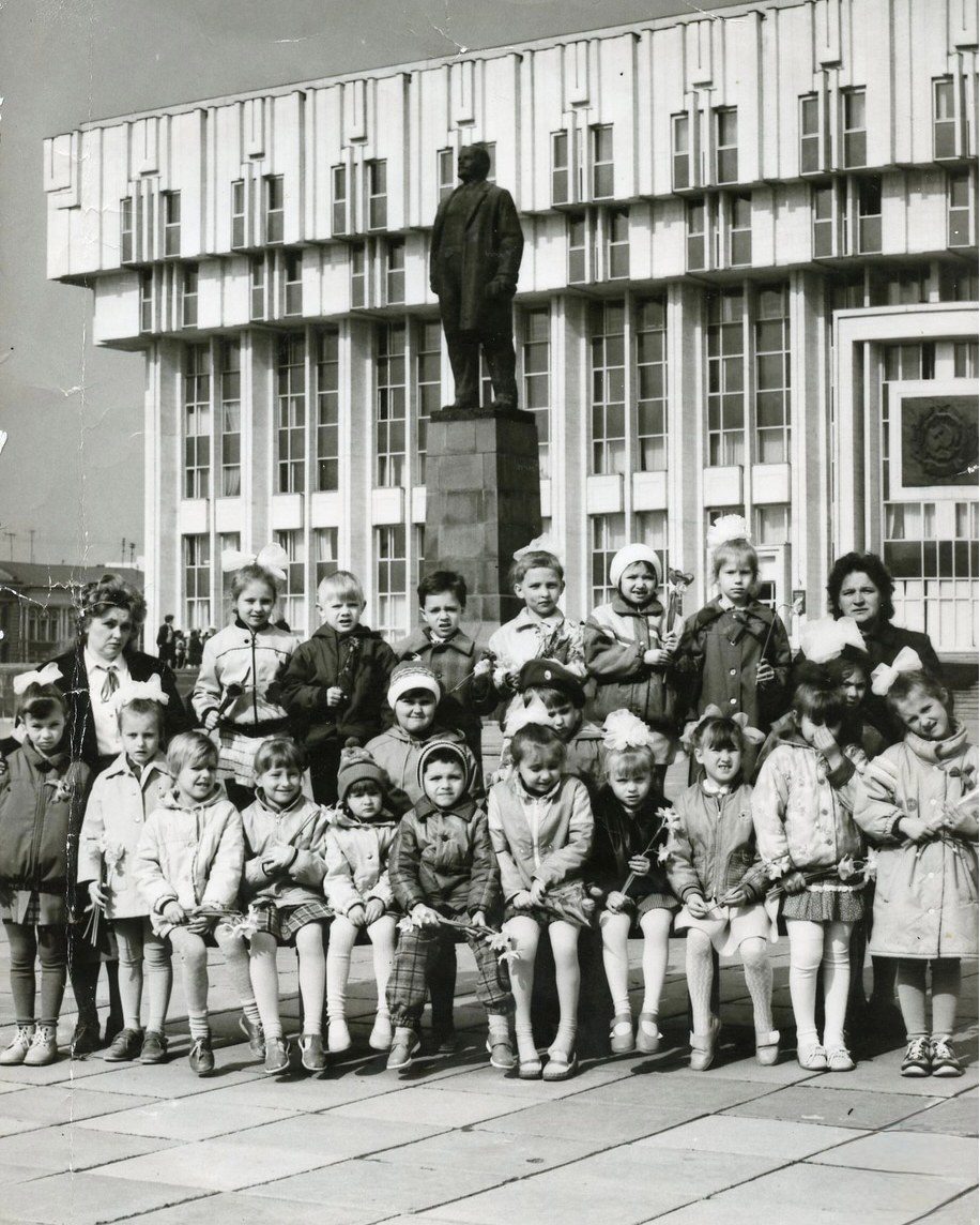 Skupinová fotografia detí a dospelých pred monumentálnou sochou a modernou budovou zo socialistickej éry.
