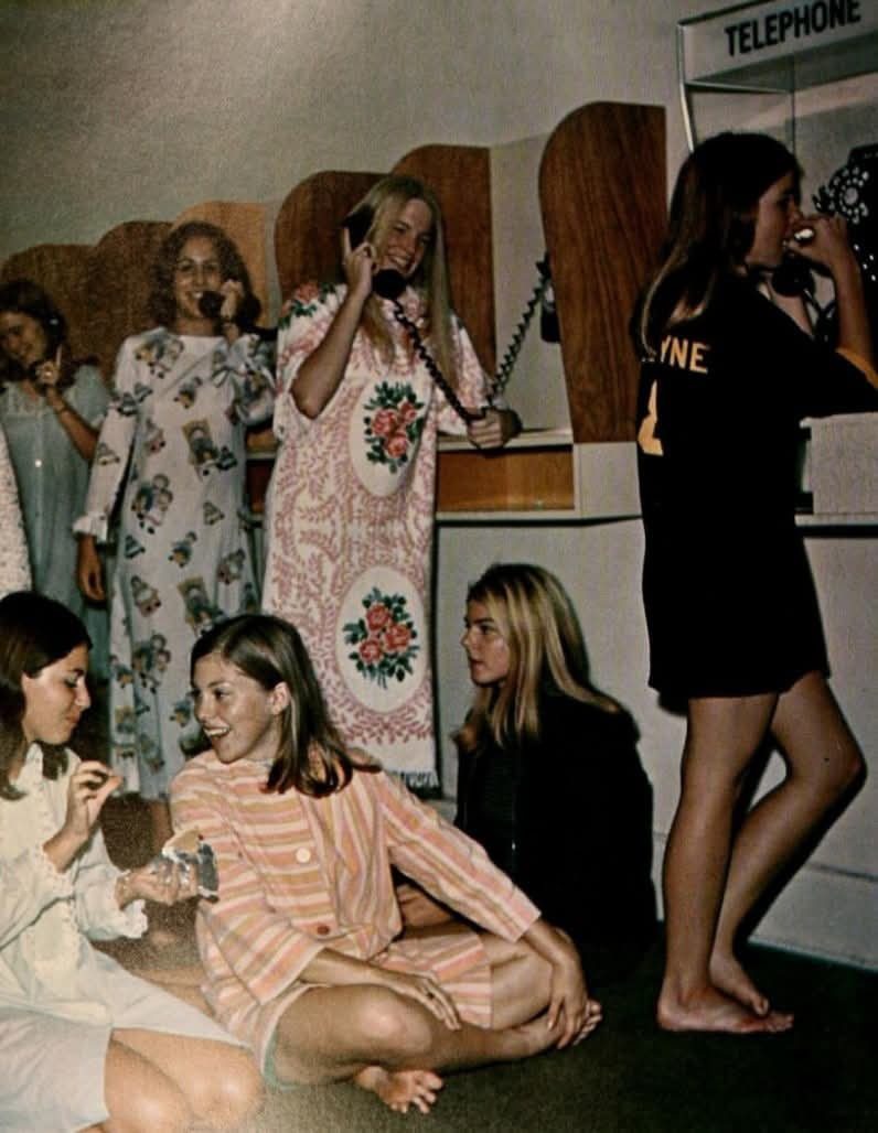 Skupina žien v pyžamách komunikuje cez staré telefóny na internáte, 1970-te roky.