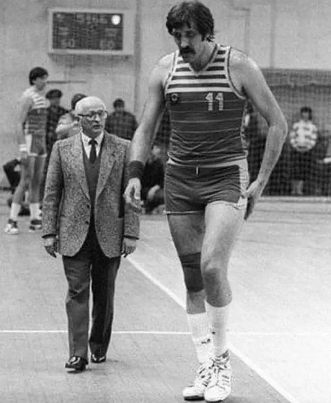 Basktetbalista v dresu s číslom 11 na ihrisku vedľa muža v obleku, čiernobiela fotografia z haly.