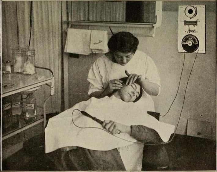 Žena v kozmetickom salóne dostáva elektrickú ošetrovaciu procedúru tváre, vintage čiernobiela fotografia.