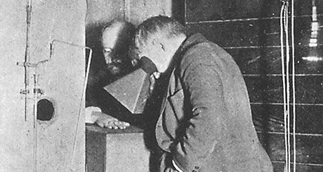 Muž počas skúmania záhadného zariadenia v čiernobielej fotografii, s historickým kontextom výzkumu alebo experimentu.