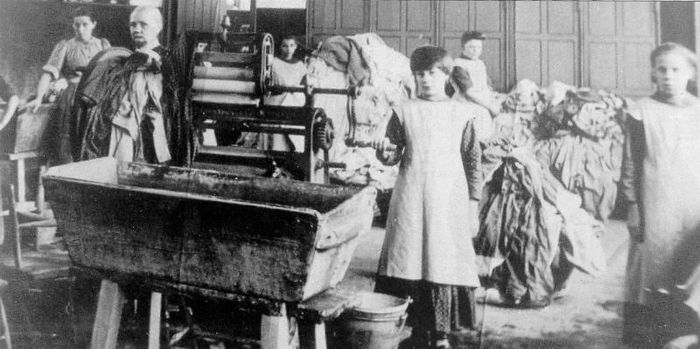 História práčovne: ženy a deti pracujú pri veľkom pracom stroji obklopené hromadami bielizne.
