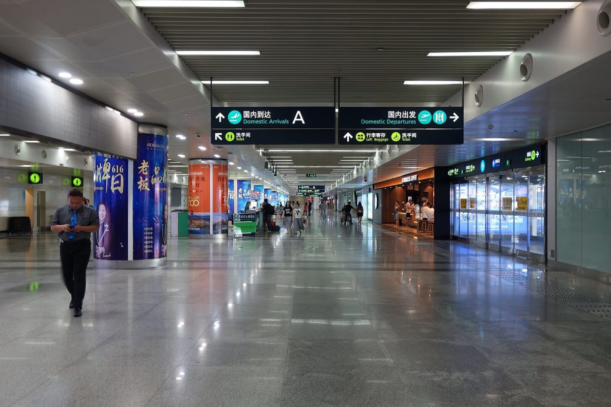 Letisková hala s cestujúcimi, smerovými tabuľami a kaviarňou. Moderný dizajn a rušná atmosféra interiéru.