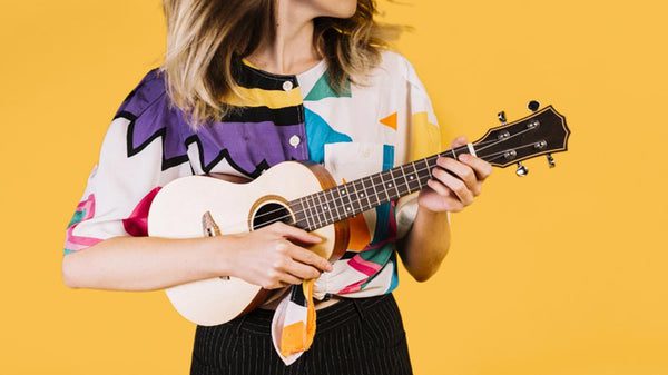 Osoba v pestrofarebnom oblečení hrá na ukulele pred žltým pozadím.