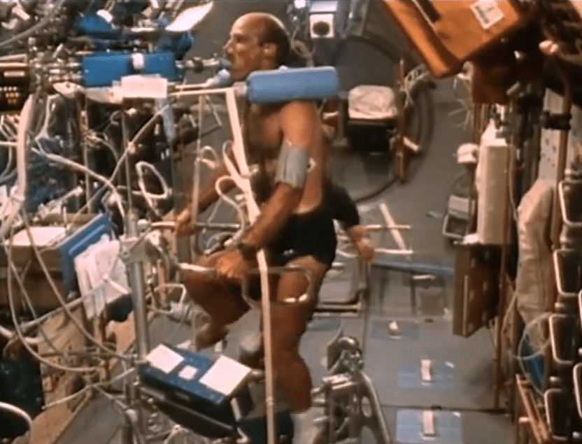 Astronaut cvičí na stacionárnom bicykli vo vesmíre, pripojený k prístrojom na monitorovanie fyziologických funkcií.