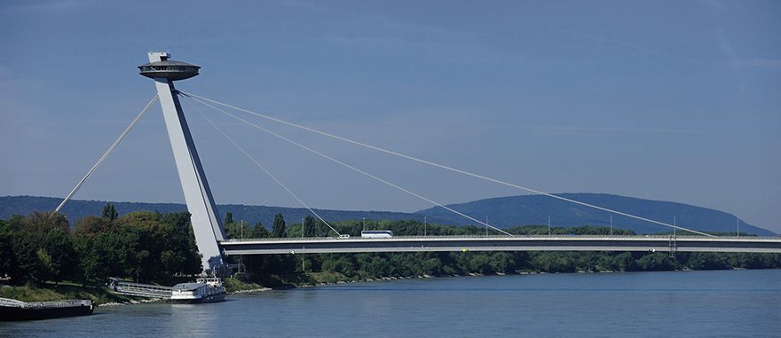 Bratislavský Most SNP cez Dunaj s vyhliadkovou vežou, modrá obloha a zelené pobrežie v pozadí.
