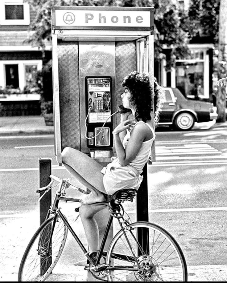 Žena sediaca na bicykli telefonuje z verejného telefónneho automatu na ulici.