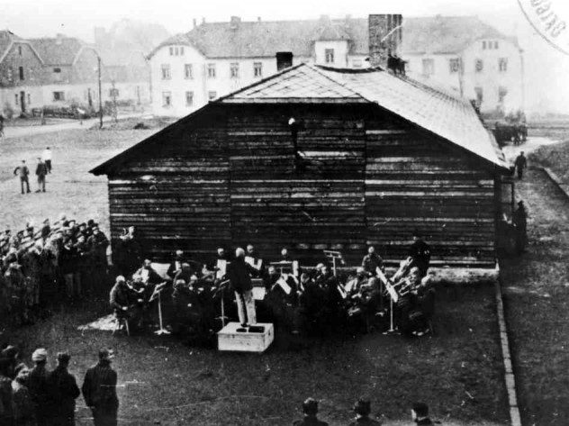 Orchester hrá koncert pred drevenou budovou, obklopený skupinou divákov v historickom mestskom prostredí.
