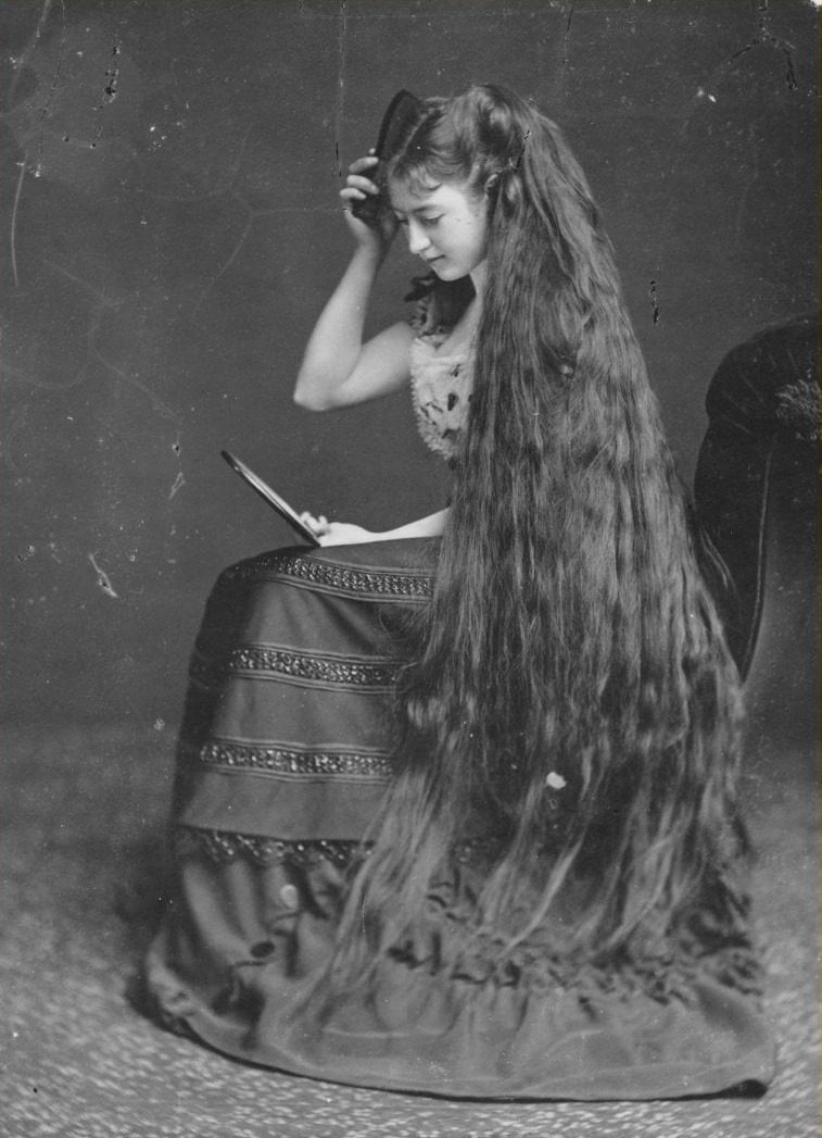 Žena s dlhými vlasmi sedí, česajúc sa hrebeňom a pozerá sa do zrkadla, historický portrét.