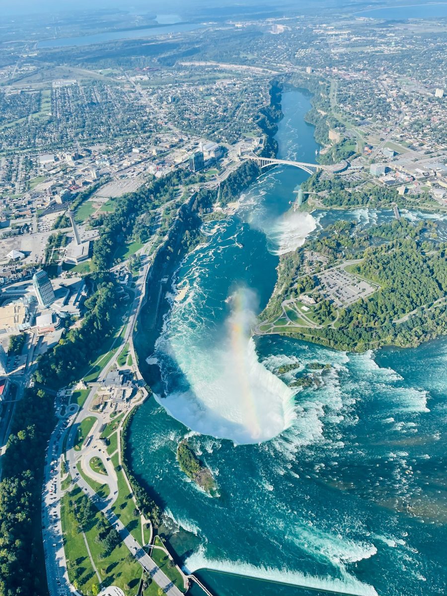 Pohľad z vtáčej perspektívy na Niagarské vodopády s dúhou, mestom a zelenými parkami v okolí.
