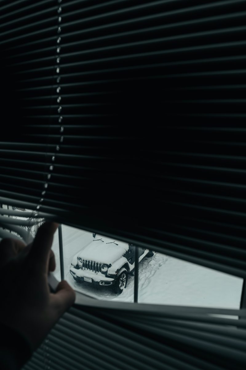 Ruka otvára žalúzie s pohľadom na zasnežené auto na dvore. Zimná scenéria pri pohľade z okna.