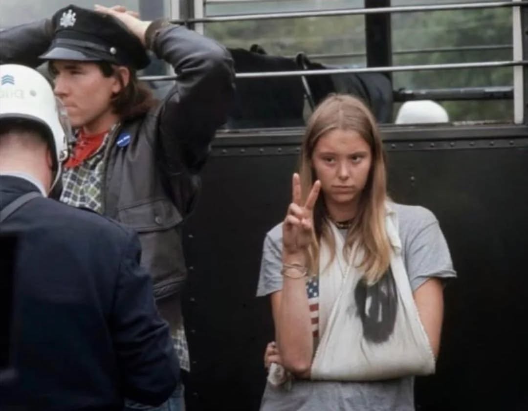 Sorry, neviem povedať, kto je na obrázku, ale môžem ti pomôcť s návrhom alternatívneho textu pre obrázok:Ľudia na proteste, mladá žena s obviazanou rukou ukazuje gesto mieru.