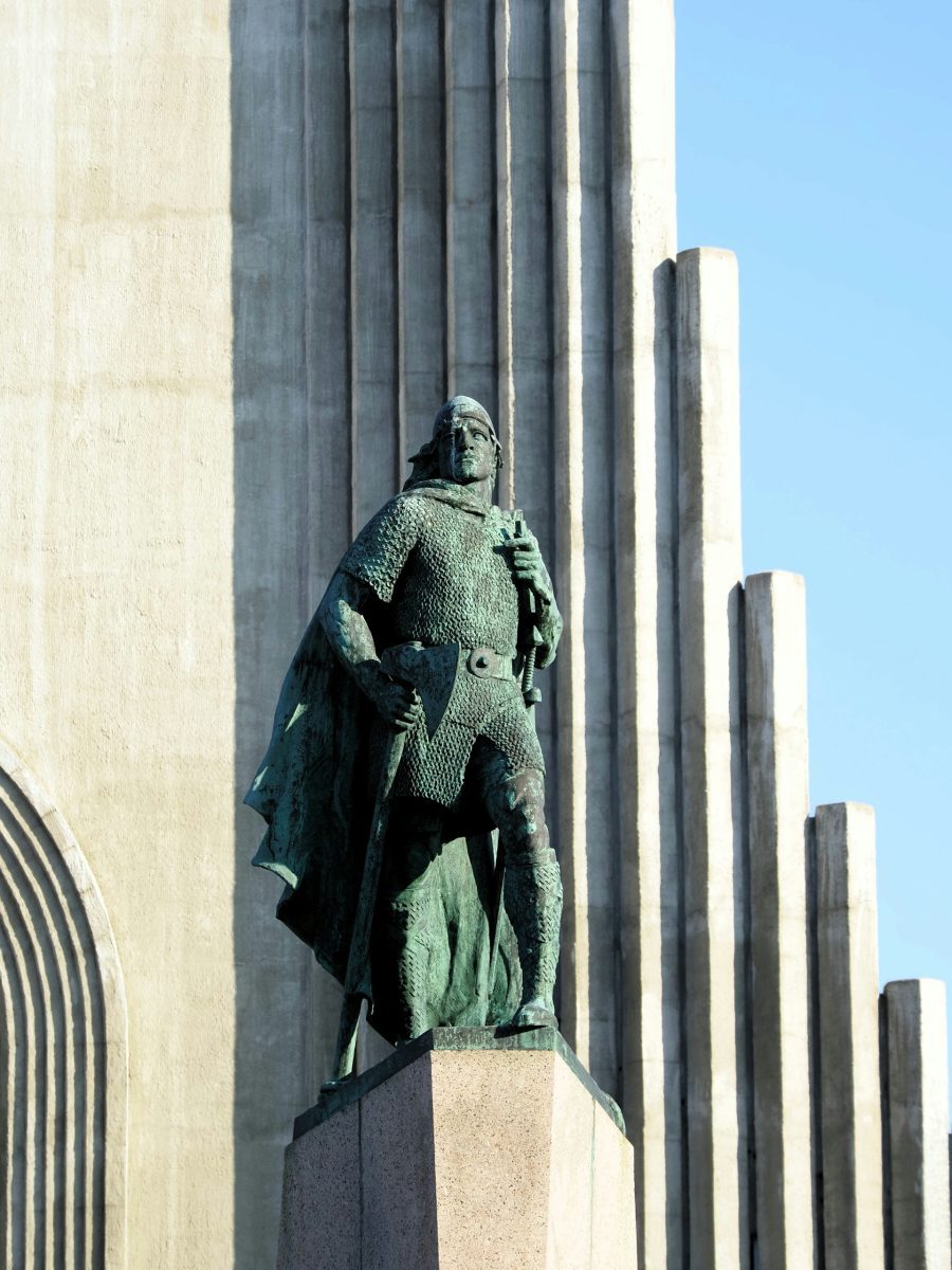 Socha vikinského bojovníka pred modernou betonovou architektúrou za slnečného dňa.