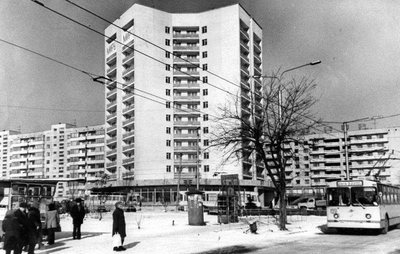 Čiernobiela fotografia panelových domov v zime s trolejbusom a ľuďmi v popredí.