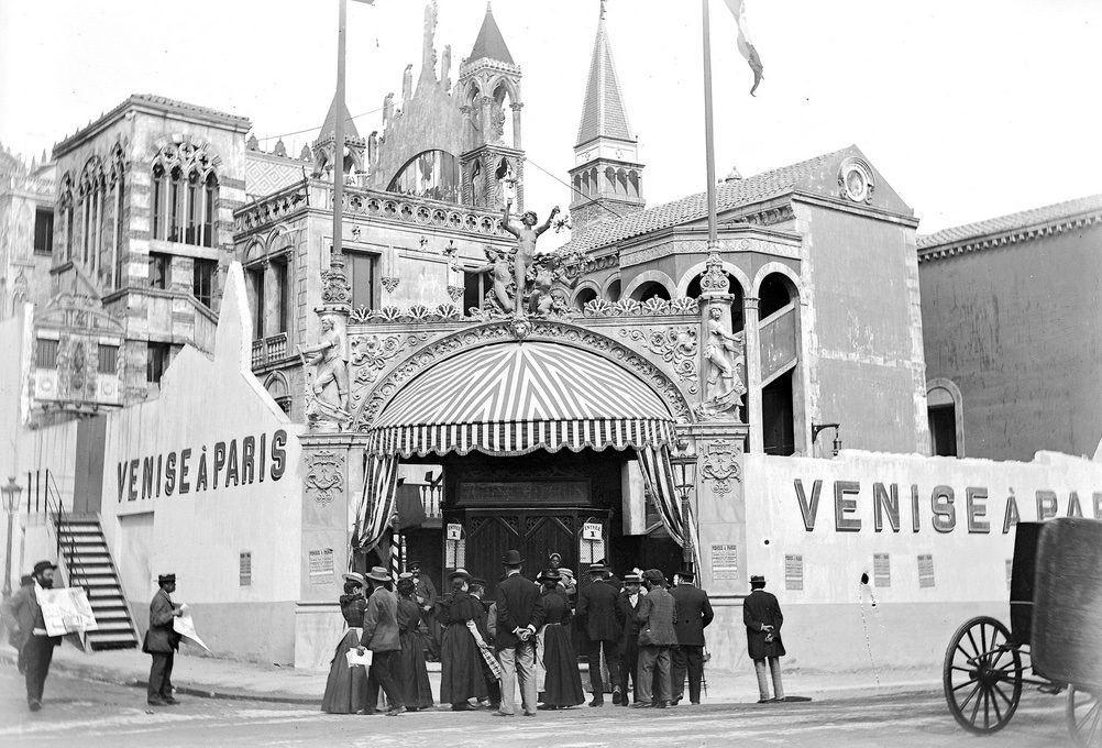 Skupina ľudí pred historickou budovou s nápismi VENISE A PARIS, zdobenou ornamentami a sochami.