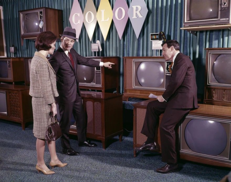 Retro obchod s farebnými televízormi, traja ľudia diskutujú v oblekoch pred výstavným panelom COLOR.