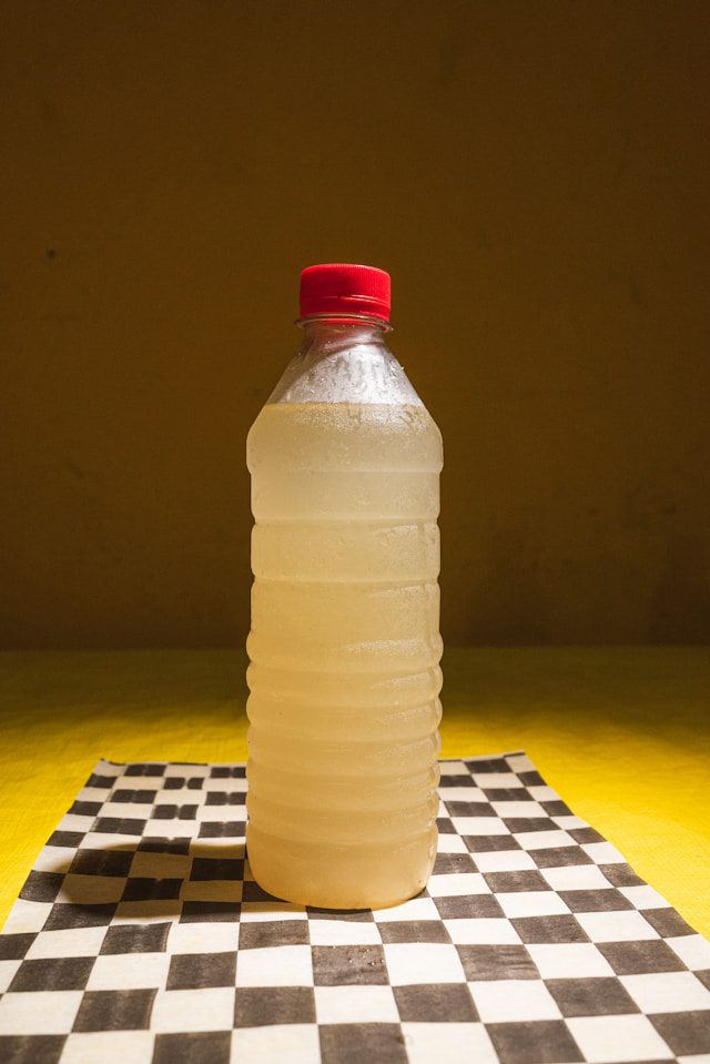 Plastová fľaša s červeným uzáverom na čierno-bielej šachovnicovej podložke, obsahujúca svetlú tekutinu.