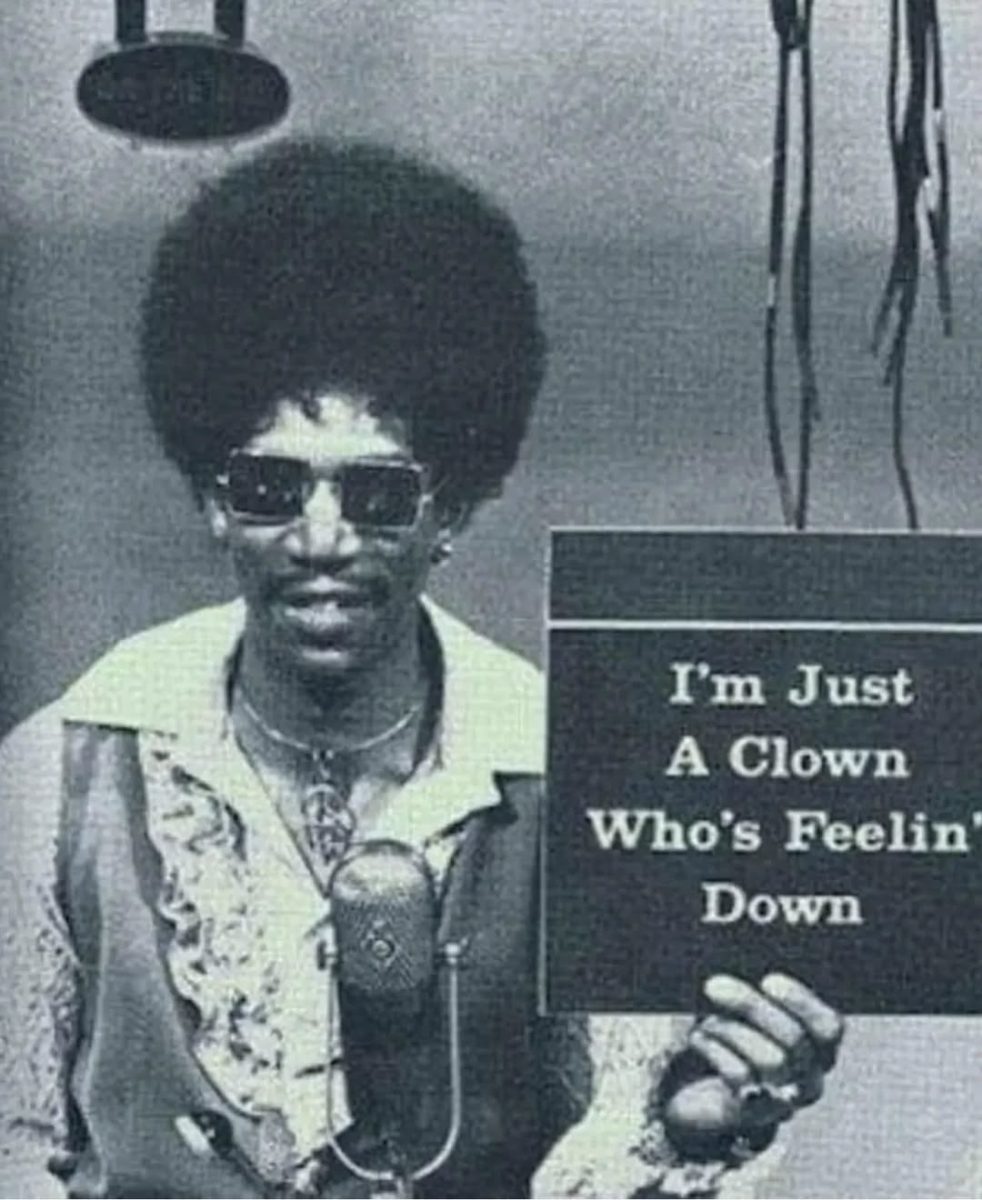 Osoba s afro účesom a okuliarmi drží tabuľu s nápisom I'm Just A Clown Who's Feelin' Down.
