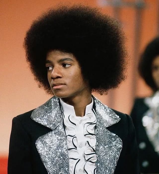 Osoba s veľkým afro účesom v elegantnom obleku so striebornými ozdobami, pozerajúca do strany.