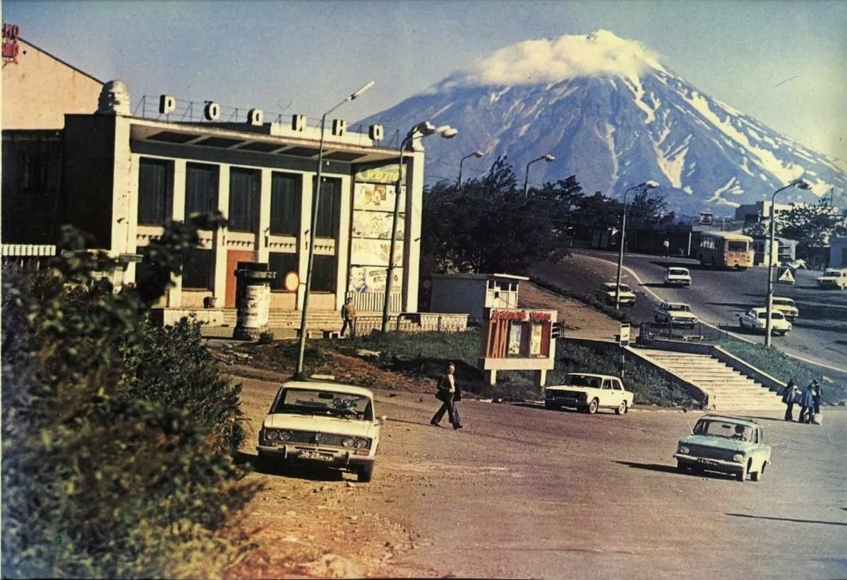 Mesto s autom na ulici, v pozadí zasnežená sopka v letnom počasí, historická atmosféra.