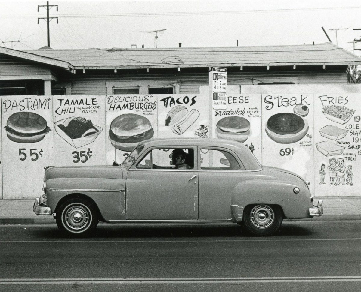 Staré auto jazdí popri stene s reklamami na jedlo, vrátane sendvičov a tacos, v vintage štýle čiernobielej fotografie.