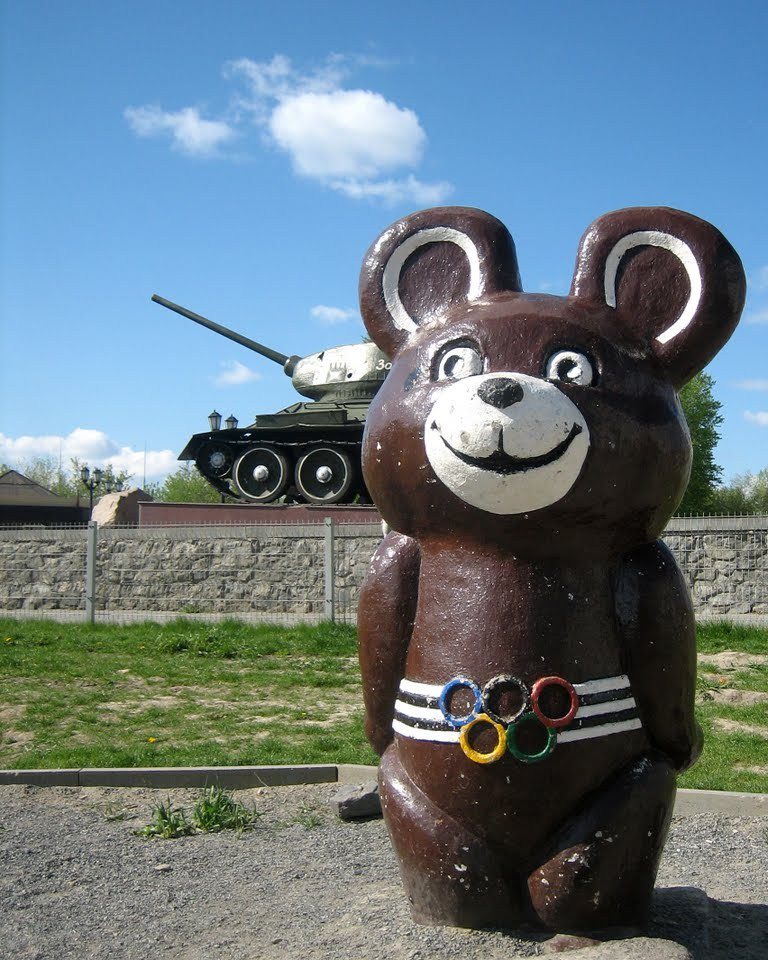 Socha medvedíka s olympijskými kruhmi, v pozadí tank, jasná obloha.