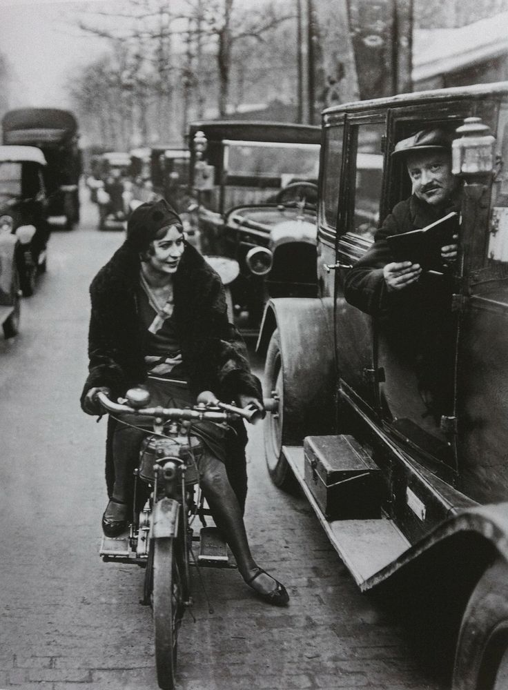 Žena na motocykli v dobovom oblečení sa usmieva na muža v aute na rušnej ulici. Historická doprava v meste.
