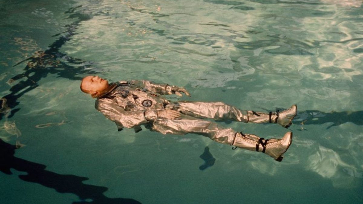 Astronaut v striebornej kombinéze pláva na hladine bazéna vo výcvikovom centre.