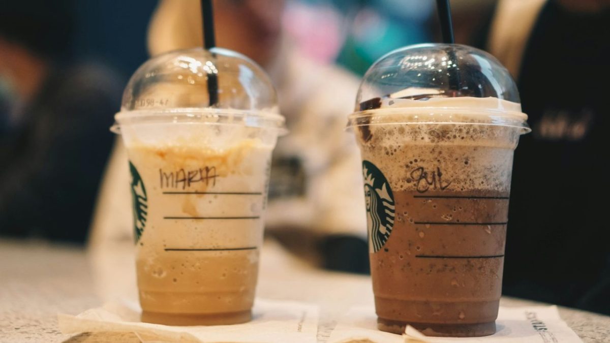 Dva ledové nápoje Starbucks na stole s plastovými vrchnákmi a slamkami, v pozadí rozostrené postavy.