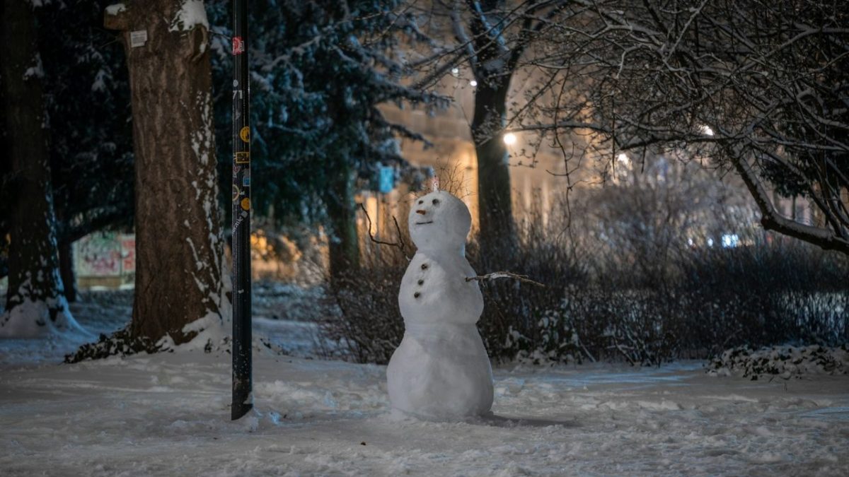 Snehuliak vo večernej zasneženej krajine pod pouličnou lampou, obklopený stromami v parku.
