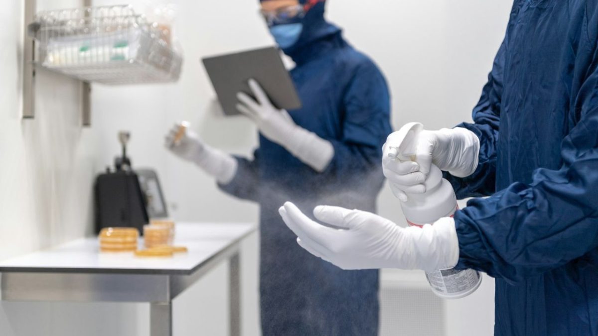 Dvaja pracovníci v ochranných odevoch dezinfikujú laboratórne prostredie pomocou spreja a tabletu.