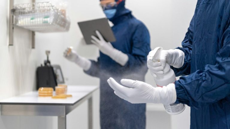Dvaja pracovníci v ochranných odevoch dezinfikujú laboratórne prostredie pomocou spreja a tabletu.
