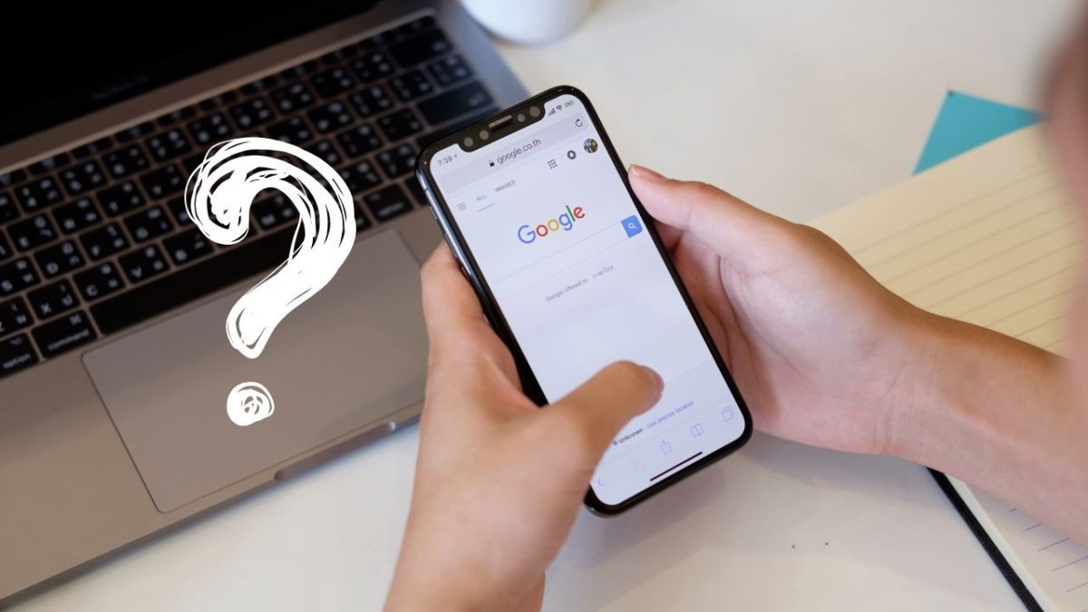Osoba hľadá niečo na Google v smartfóne pred otvoreným notebookom a otáznikom.