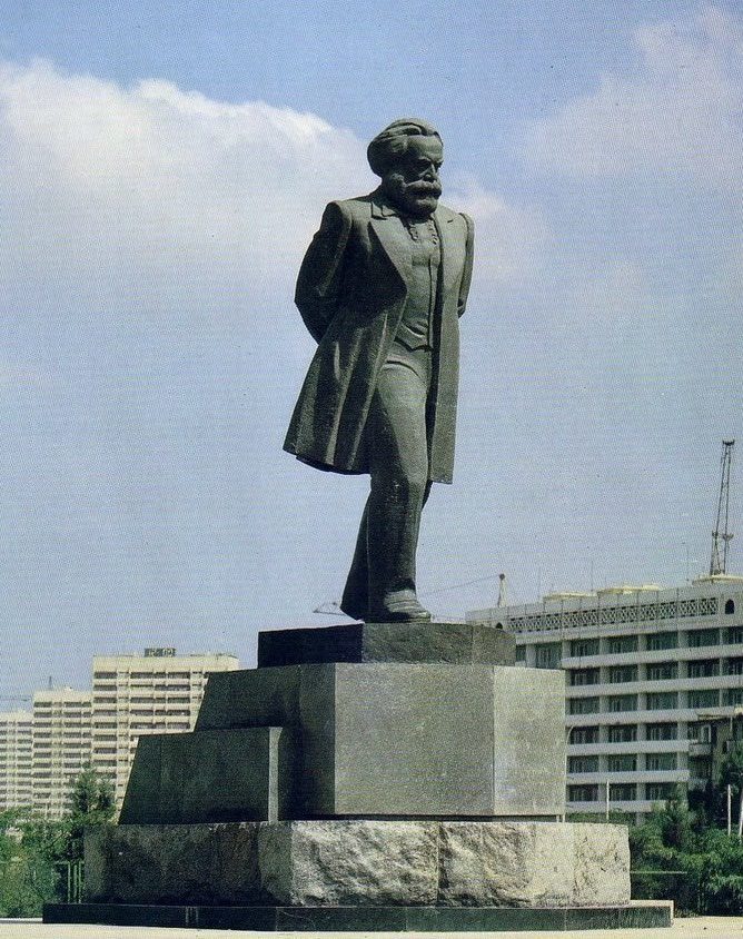 Socha postavy na podstavci v mestskom prostredí, obklopená budovami a modrou oblohou v pozadí.