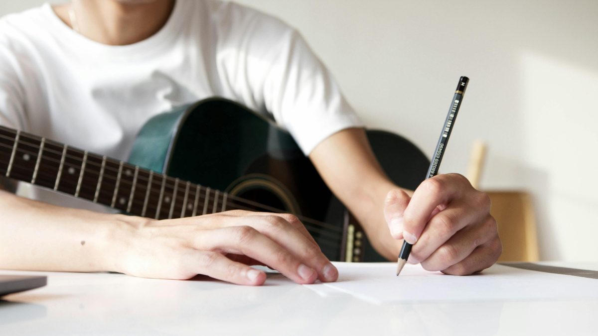 Osoba s gitarou píše na papier, zachytená v momente tvorby hudby alebo písania textov.