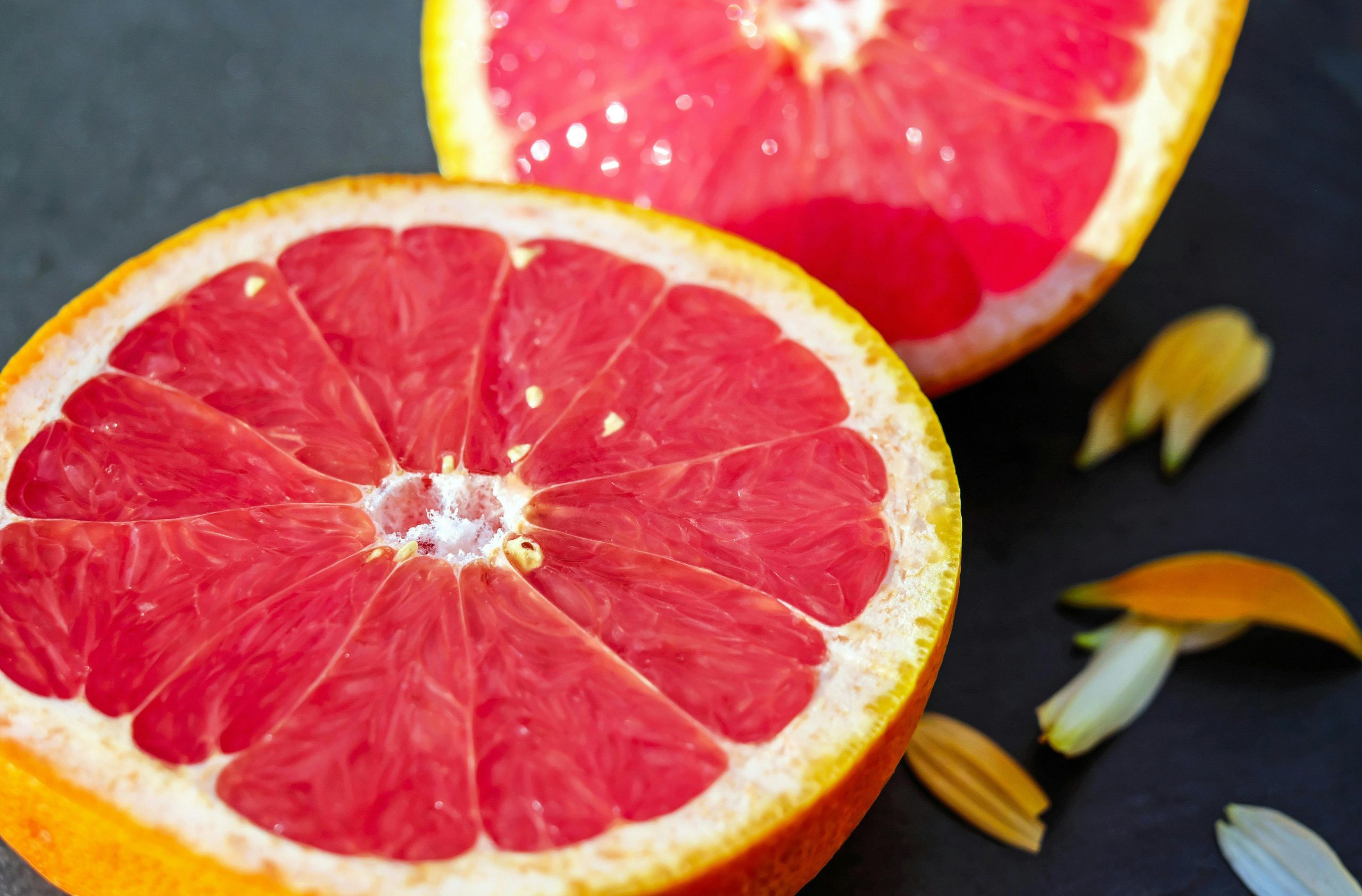Ružový grapefruit rozdelený na polovicu, na tmavom pozadí, s niekoľkými okvetnými lístkami.