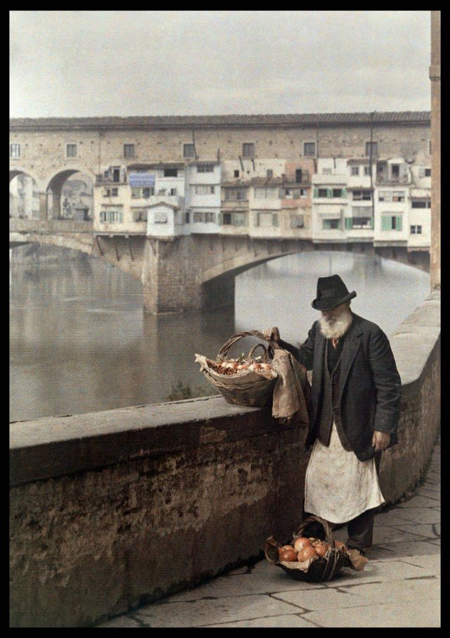 Starý muž predáva ovocie v košíkoch pri rieke Arno vo Florencii, s historickým mostom Ponte Vecchio v pozadí.
