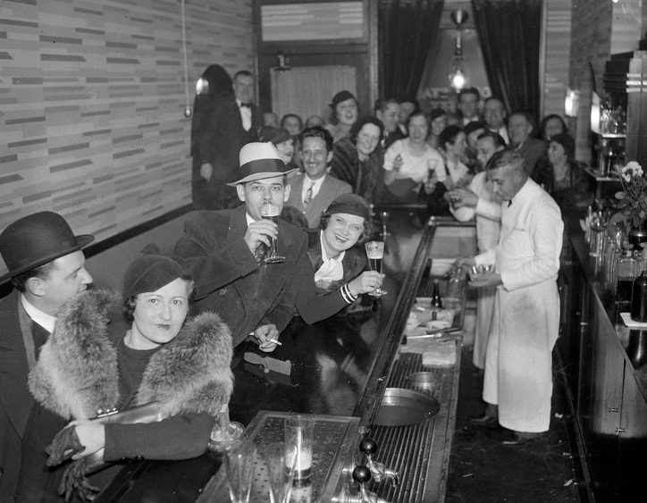 Hostia v štýlovom bare pijúci nápoje, barman pripravuje kokteily, živá atmosféra 1930-tych rokov.