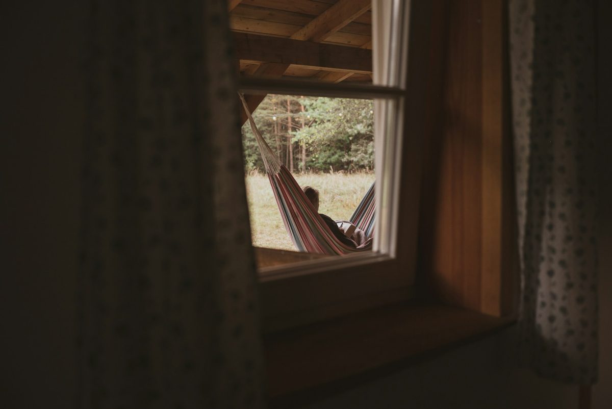 Osoba relaxujúca v hamake na verande, pohľad cez okno, les v pozadí.