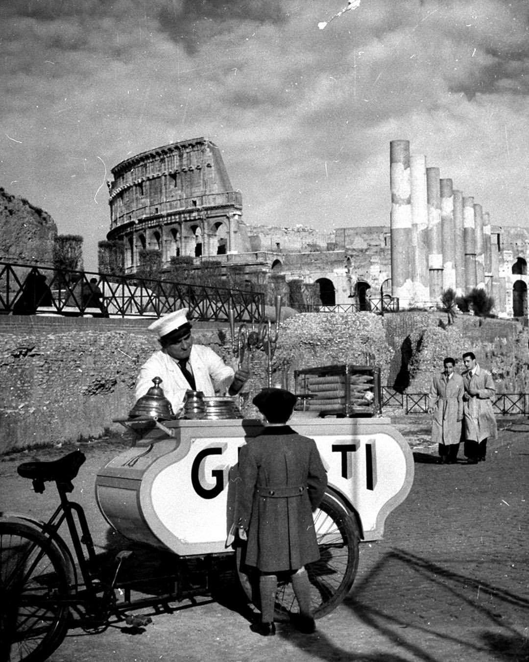 Predajca zmrzliny a dieťa pri košíku s nápisom Gelati pred Koloseom v Ríme, historická čiernobiela fotografia.