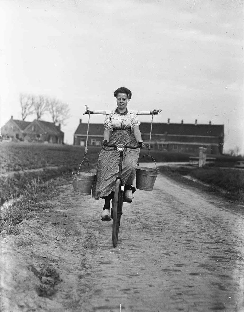 Žena na bicykli vezie vedrá po vidieckej prašnej ceste, starobylá dedina v pozadí.