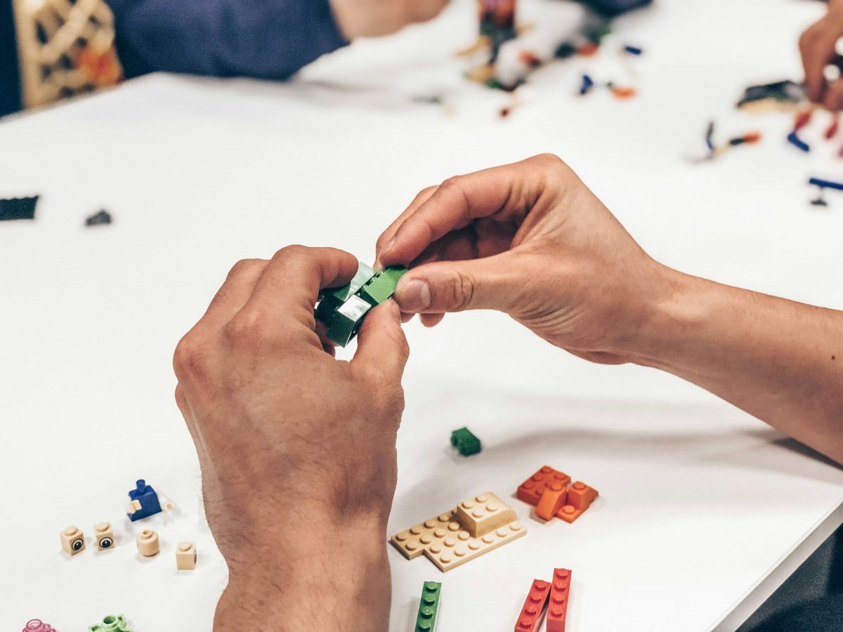 Ruky skladajú rôznofarebné LEGO kocky na bielom stole, detailný záber na kreativitu.