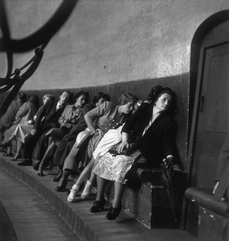 Ženy sediace na lavici, opierajúce sa o stenu, zvnútra miestnosti. Historická atmosféra, čiernobiela fotografia.