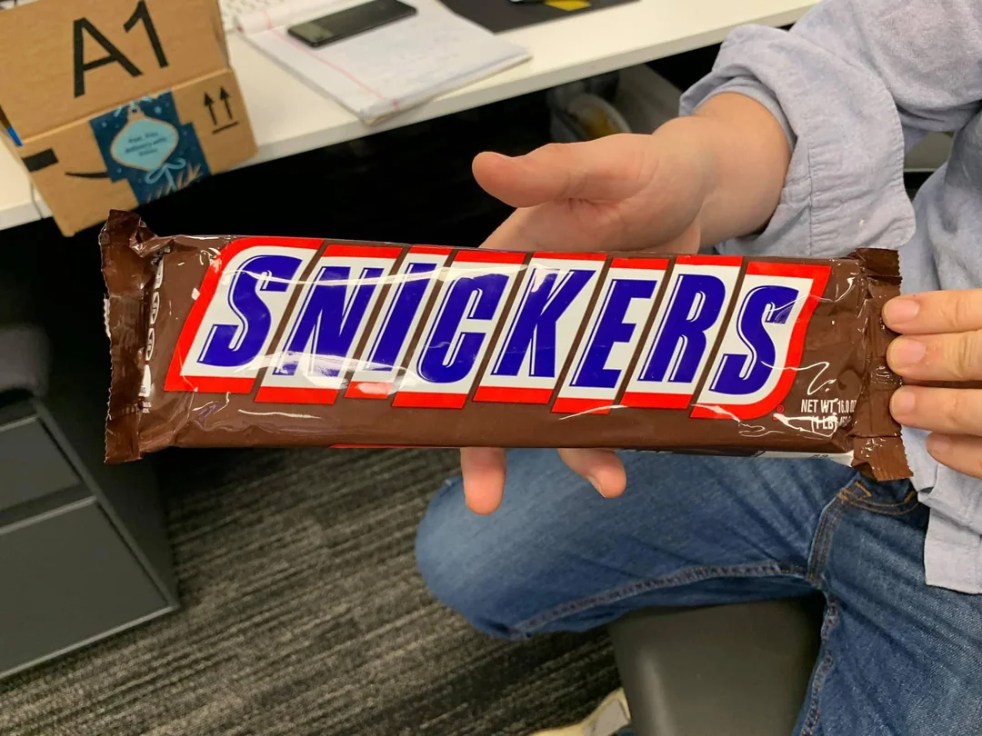Osoba drží veľkú čokoládovú tyčinku Snickers na pracovnom stole s balíkom v pozadí.