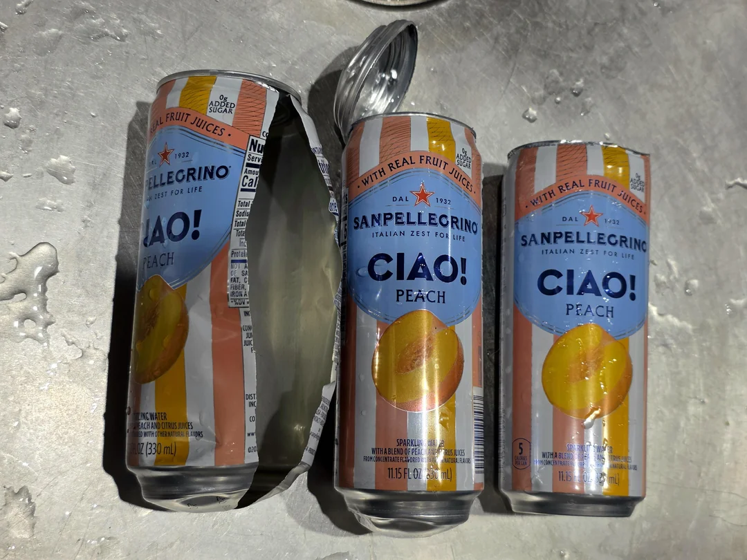 Tri plechovky Sanpellegrino Ciao! Peach na mokrom povrchu, jedna otvorená.