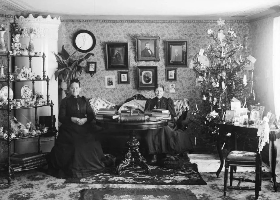Dve ženy v historickom interiéri s vianočným stromčekom a starožitnými dekormi, čiernobiela fotografia.