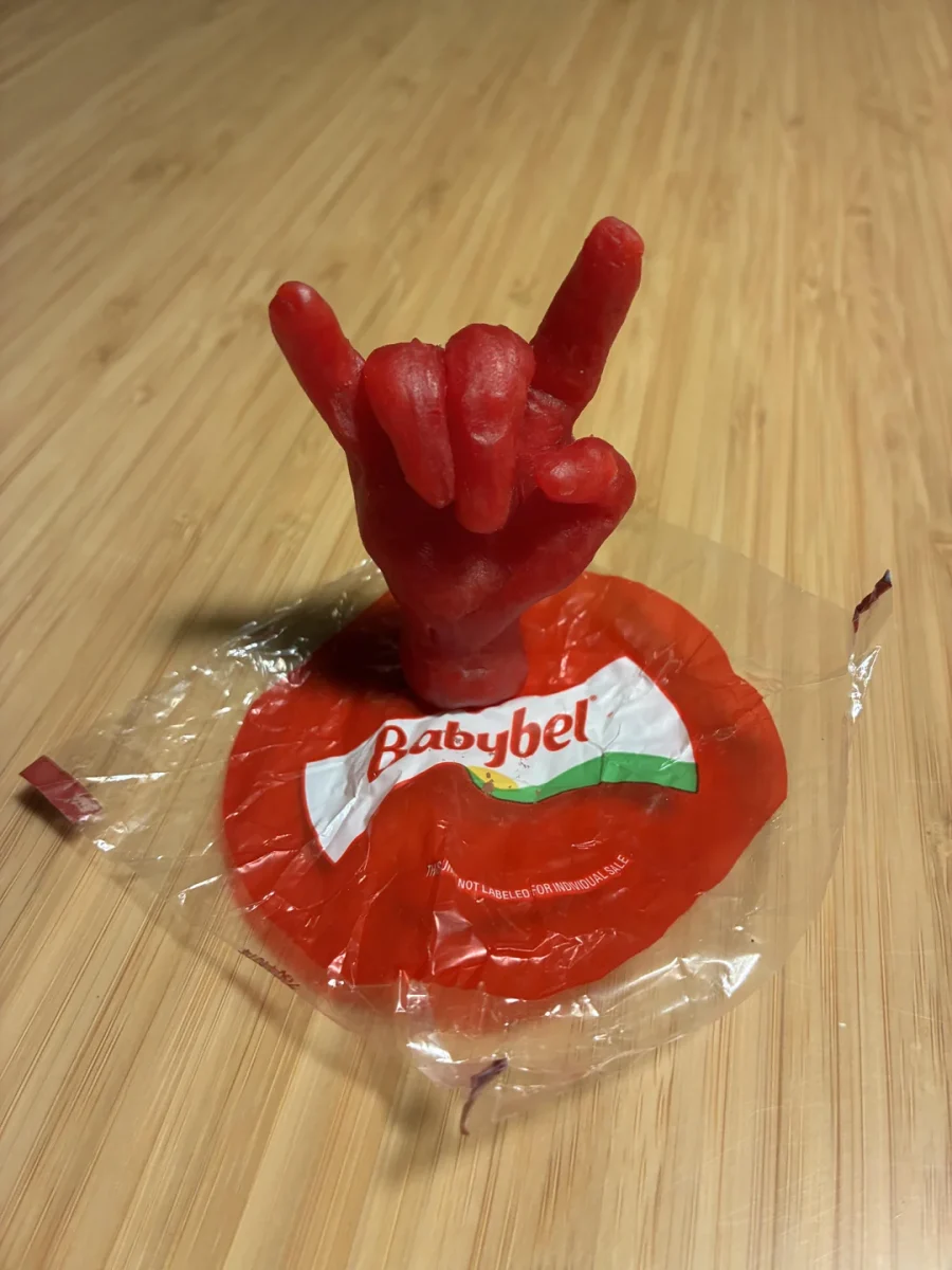 Červený voskový obal Babybel syra v tvare ruky s rockovým gestom na drevenom stole.